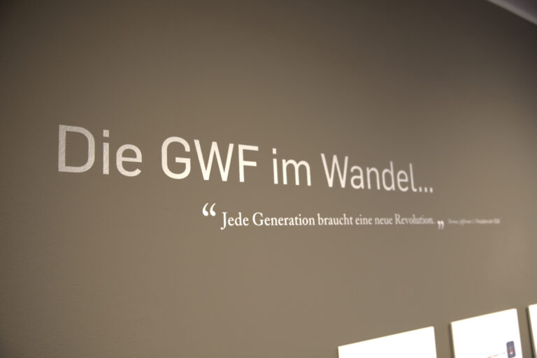 Foto. Dient der visuellen Einleitung eines Ausstellungsbereichs zum Wandel der Logos der GWF. Wand mit großflächigem Schriftzug „Die GWF im Wandel…“. Darunter Zitat: „Jede Generation braucht eine neue Revolution.“. Neutral beleuchtete, glatte Wandfläche, rechts unten beginnt eine Reihe gerahmter Logos. Quelle: Laura Angelina Wörner/THWS