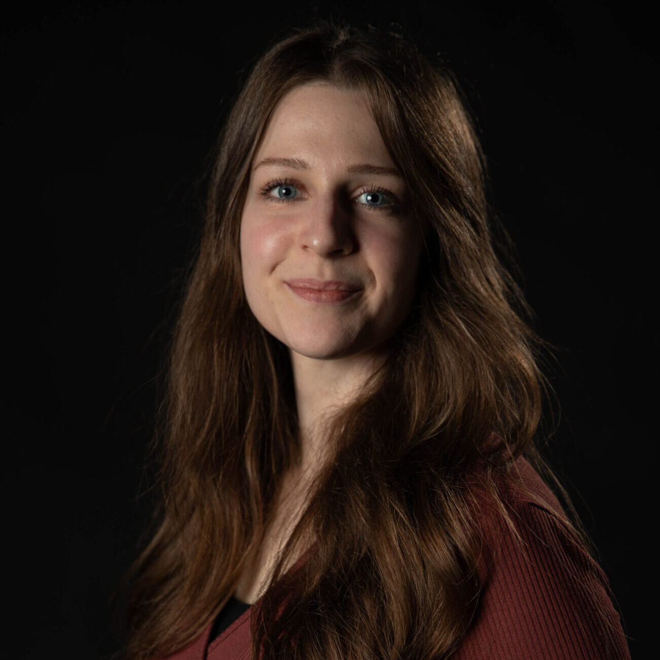 Portrait. Dient der Vorstellung der genannten Person. Eine junge Frau steht vor einem schwarzen Hintergrund. Sie trägt ein dunkelrotes Oberteil. Die linke Schulter ist leicht nach vorn gedreht, sie schaut gerade und freundlich lächelnd in die Kamera. Quelle: Lynn Emmerling