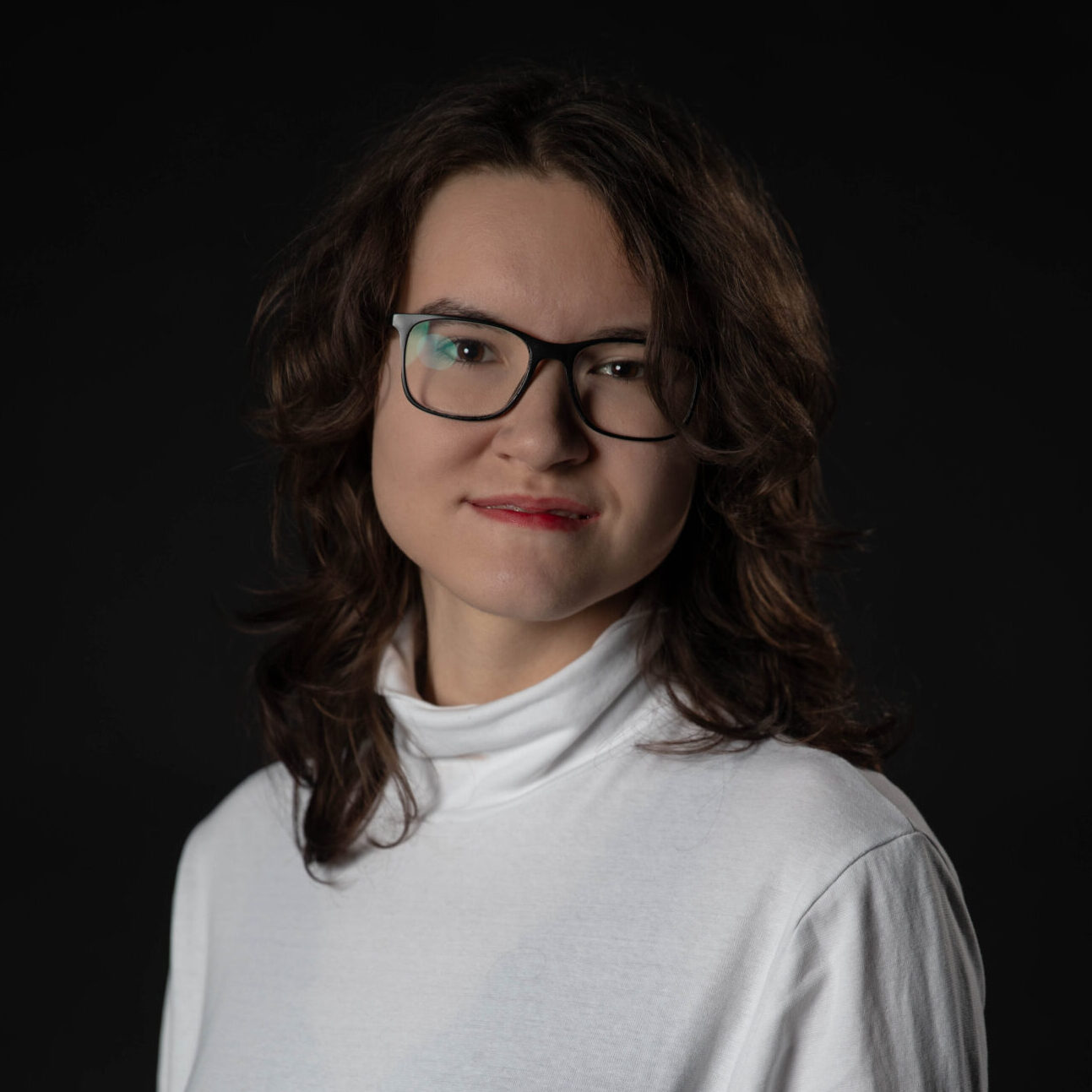 Portrait. Dient der Vorstellung der genannten Person. Eine junge Frau steht vor einem schwarzen Hintergrund. Sie trägt ein weißes Oberteil. Die linke Schulter ist leicht nach vorn gedreht, sie schaut gerade und freundlich lächelnd in die Kamera. Quelle: Angelina Zelch