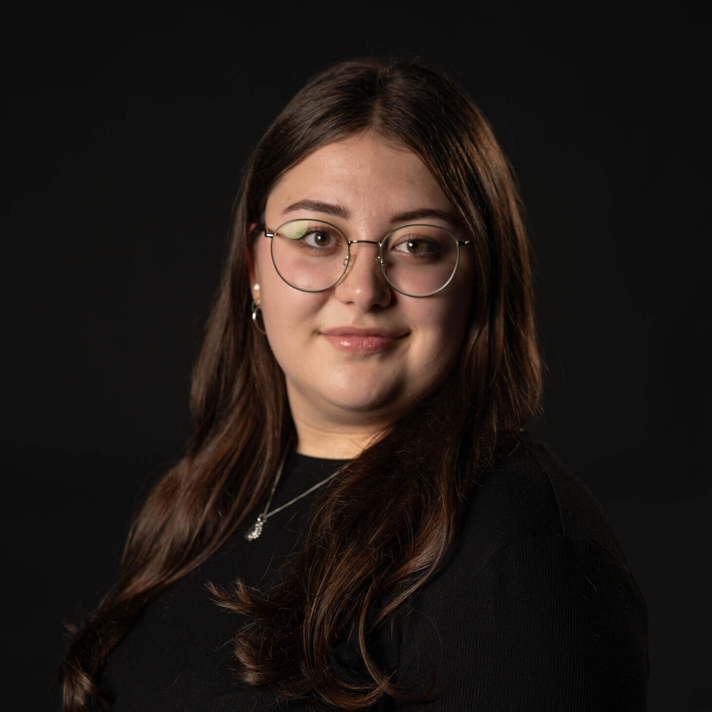 Portrait. Dient der Vorstellung der genannten Person. Eine junge Frau steht vor einem schwarzen Hintergrund. Sie trägt ein schwarzes Oberteil. Die linke Schulter ist leicht nach vorn gedreht, sie schaut gerade und freundlich lächelnd in die Kamera. Quelle: Julia Soppa